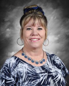 A smiling woman with blonde hair styled in a bun, wearing a patterned top and a blue flower necklace.