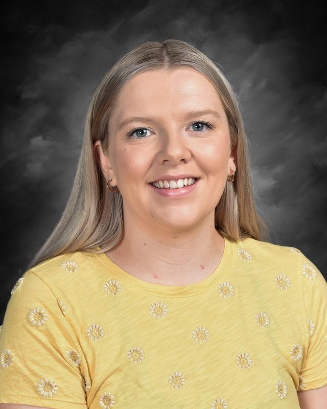 Smiling woman with long blonde hair wearing a yellow shirt with floral designs, against a dark background.