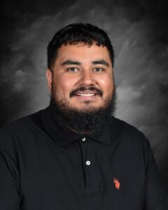 A man with a beard smiles at the camera, wearing a black collared shirt with a logo, against a dark background.