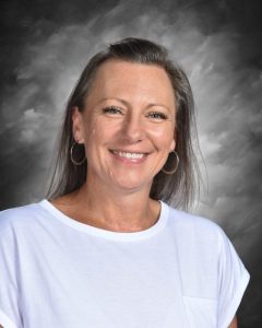 Mujer sonriente con pendientes de aro y una camisa blanca sobre un fondo gris nublado.