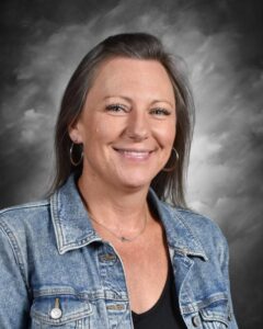 A smiling woman with long hair wearing a denim jacket and hoop earrings, set against a cloudy background.