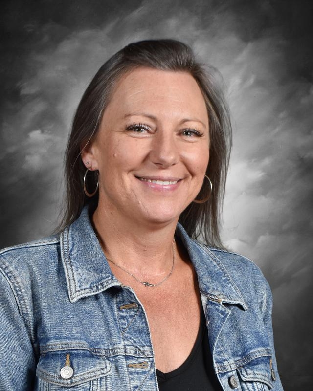A smiling woman with long hair wearing a denim jacket and hoop earrings, set against a cloudy background.