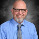 A smiling man with glasses and a neatly trimmed beard, wearing a blue shirt and gray tie, set against a gray background.