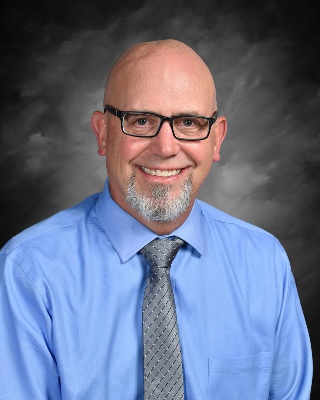 A smiling man with glasses and a neatly trimmed beard, wearing a blue shirt and gray tie, set against a gray background.