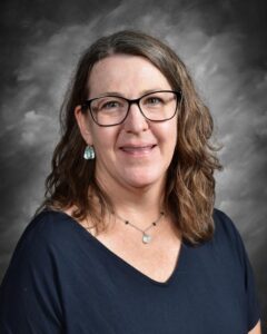 A smiling woman with shoulder-length brown hair, wearing glasses and a dark shirt, against a gray background.