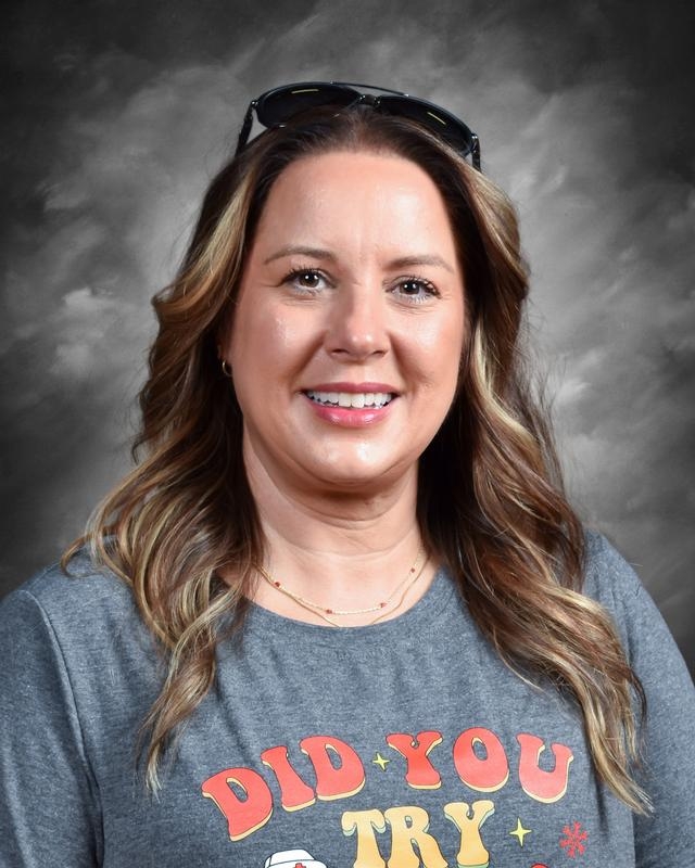 A smiling woman with long, wavy brown hair wearing a gray shirt with colorful text, and sunglasses on her head.
