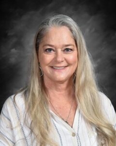 A smiling woman with long, straight gray hair, wearing a white button-up shirt with stripes, against a dark background.