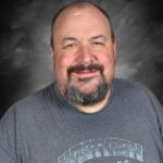 A smiling man with a beard wears a gray shirt advertising "West M Wildcats," against a soft gray background.