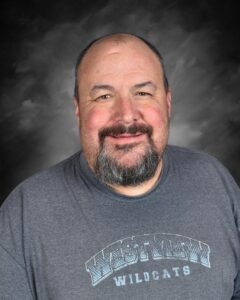 A smiling man with a beard wears a gray shirt advertising "West M Wildcats," against a soft gray background.