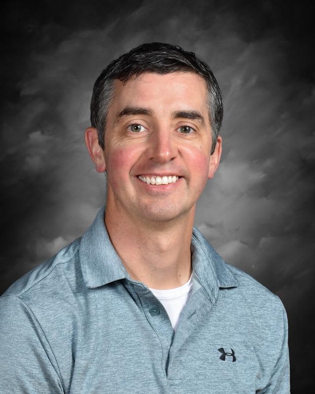 A man with short hair smiles, wearing a gray polo shirt, set against a dark, cloudy background.
