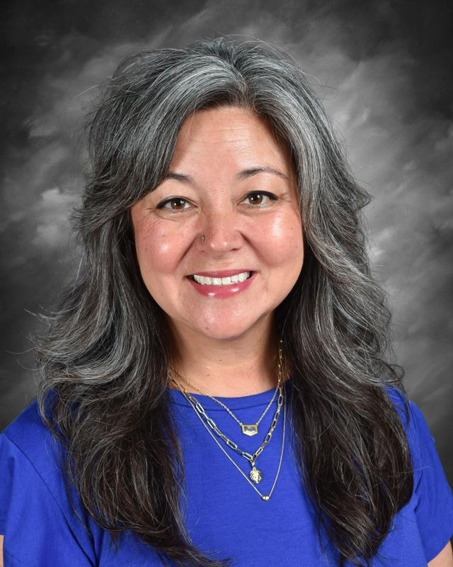 Smiling woman with long gray hair, wearing a blue shirt and layered necklaces, set against a gray background.