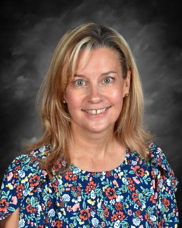 Smiling woman with blonde hair wearing a floral blouse against a dark background.