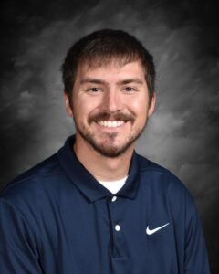 Smiling man with short dark hair and a beard, wearing a navy blue polo shirt against a gray background.