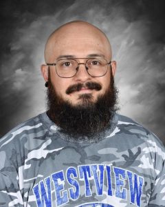 Hombre calvo, con gafas y barba, que lleva una camiseta de camuflaje con la inscripción «Westview», sobre un fondo nublado.