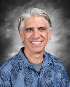 Smiling man with gray hair in a blue patterned shirt against a cloudy backdrop.