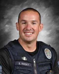 Smiling police officer in uniform with a badge, against a gray background. He has short hair and is looking directly at the camera.