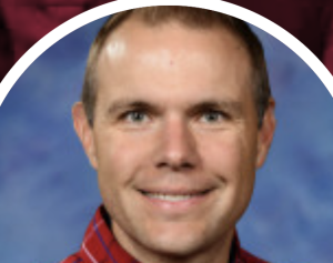 Headshot of a smiling man with short brown hair, wearing a red plaid shirt, against a blue background.