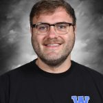 Hombre con gafas que sonríe delante de un fondo gris, vestido con una camiseta negra con el logotipo «W» azul y la inscripción «Wildcats».