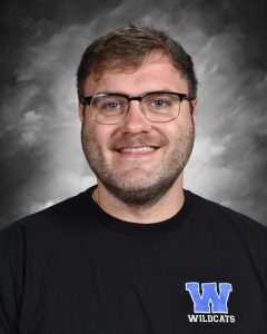 Hombre con gafas que sonríe delante de un fondo gris, vestido con una camiseta negra con el logotipo «W» azul y la inscripción «Wildcats».