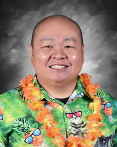 Smiling man wearing a colorful Hawaiian shirt with a cat design and a floral lei, against a gray backdrop.