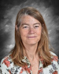 A woman with long hair and a floral blouse smiles against a gray background.