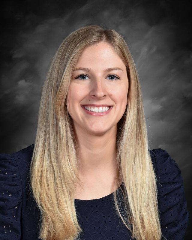 Smiling woman with long blonde hair, wearing a navy blouse, against a dark, cloudy background.