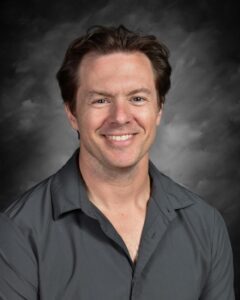 Smiling man with short hair wearing a gray shirt, set against a cloudy gray background.
