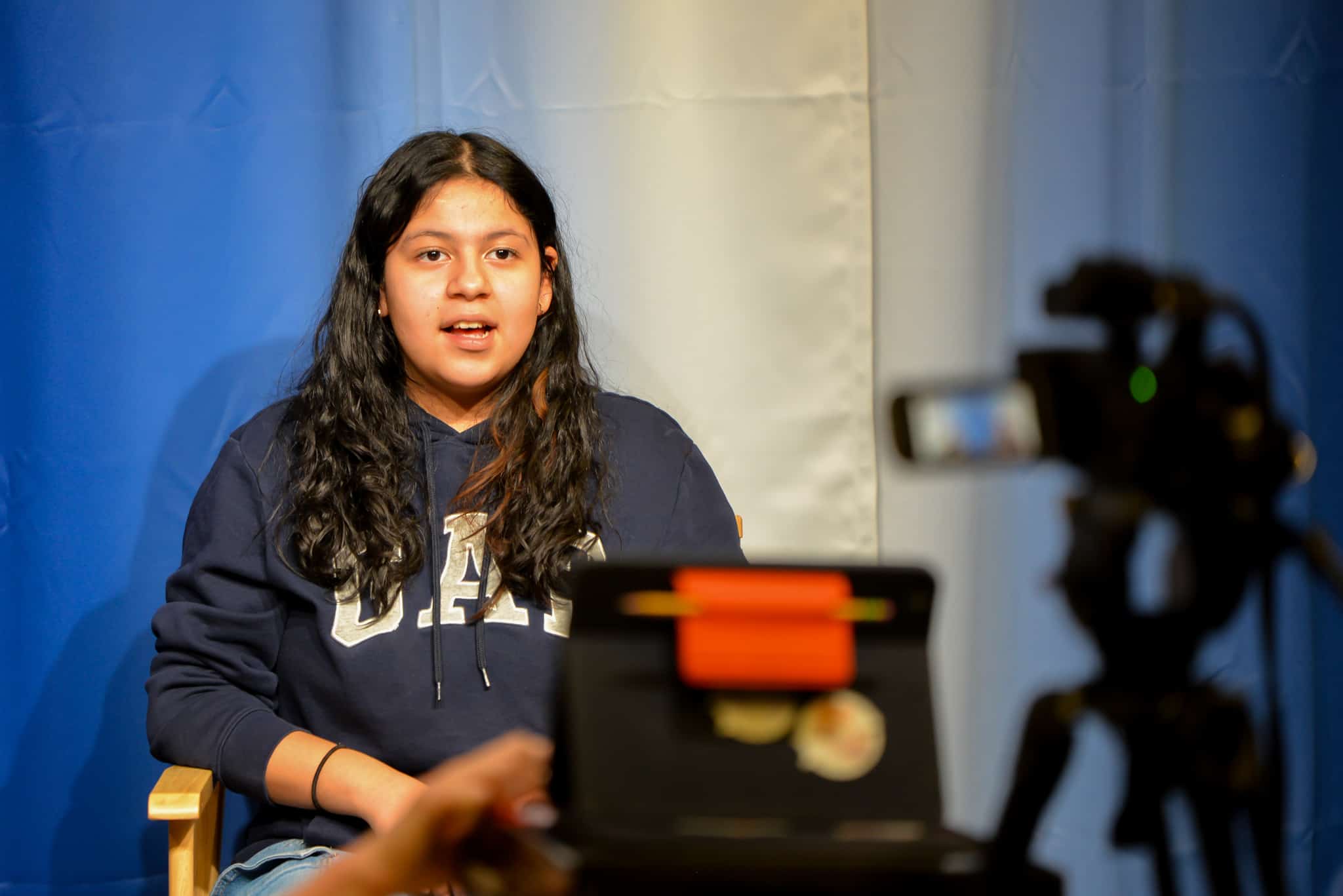 Female middle school student sitting on a chair in front of a camera. She is talking on camera during a newscast that is being recorded.
