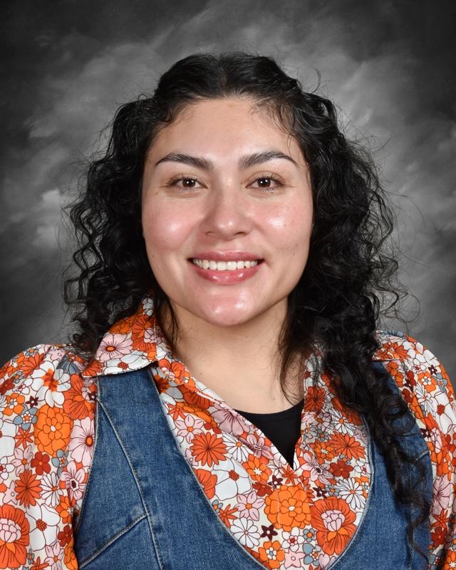 Smiling person with curly hair, wearing a floral shirt and denim vest, against a gray background.