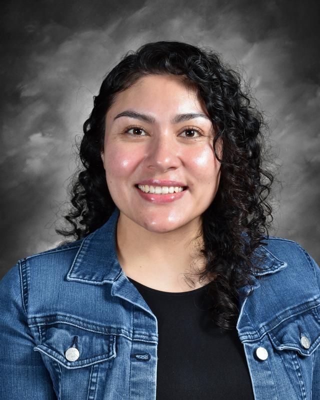 A smiling woman with curly hair wearing a denim jacket over a black top, set against a gray background.