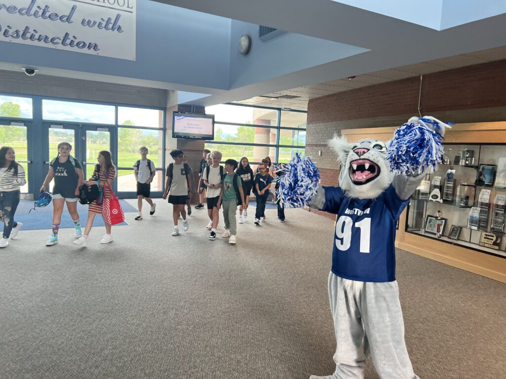 Westview Wildcat Mascot, Waffles, cheering on the right of the screen with students walking in the school behind him.