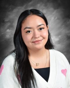 A young woman with long black hair smiles, wearing a white sweater with pink hearts against a gray background.