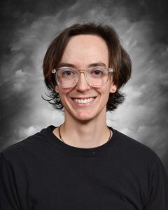 Una persona con el pelo hasta los hombros y gafas, vestida con una camiseta negra, sonríe ante un fondo gris y nublado.