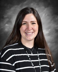 A woman with long brown hair smiles, wearing a black and white striped shirt and a necklace, against a gray backdrop.