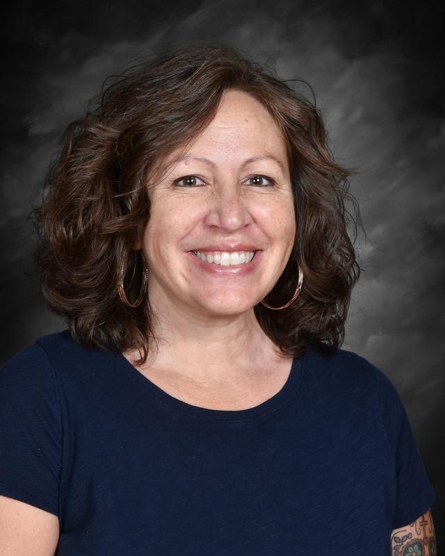 Smiling woman with curly brown hair, wearing a navy shirt and hoop earrings, against a dark gray background.