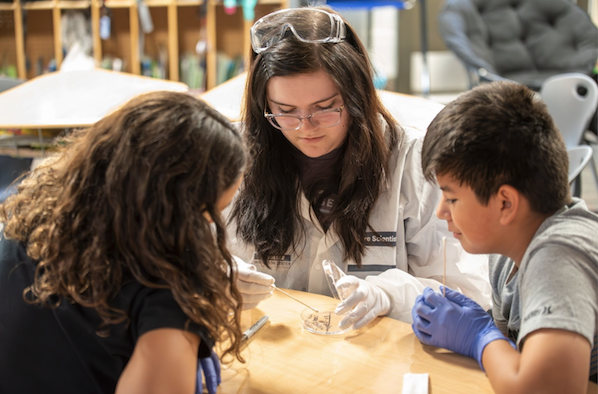A young scientist demonstrates an experiment to two children at a table filled with laboratory equipment and tools.