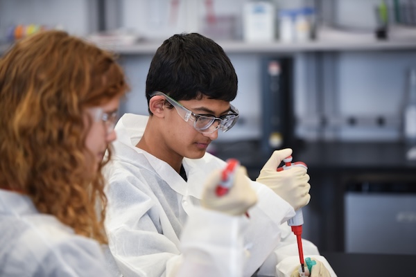 A teenager in safety goggles uses a pipette while a classmate works nearby in a laboratory setting.