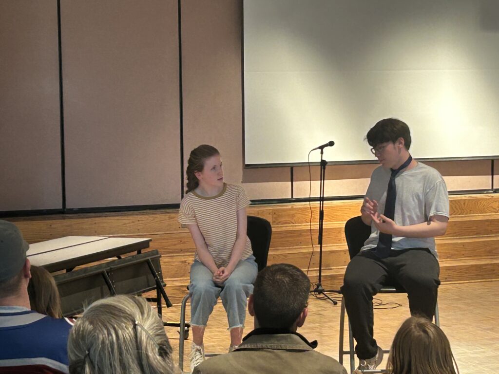 Two young people sit in chairs on stage during a discussion; one speaks while the other listens attentively.