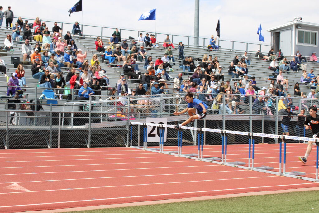 A runner in a blue uniform clears a hurdle during a track event, with spectators in the background.