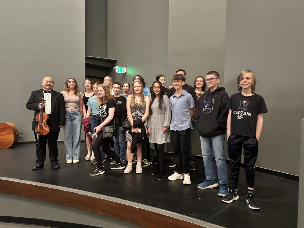 A group of students and a violinist pose together on stage after a performance.
