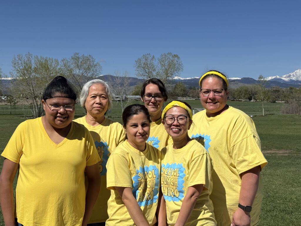 A group of six individuals wearing yellow shirts poses outdoors with mountains in the background on a sunny day.