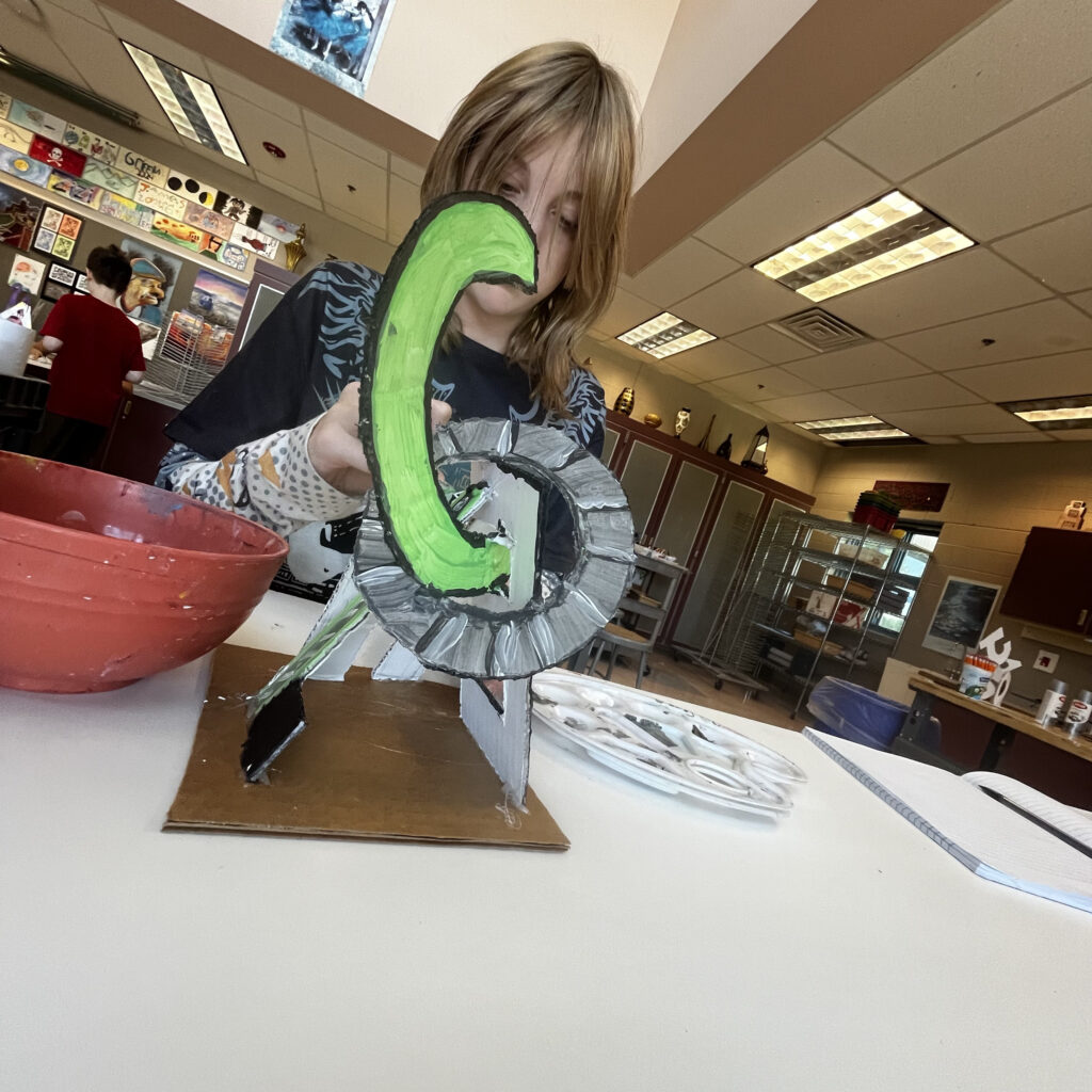 A child is painting a colorful sculpture in an art classroom, with various art supplies and projects in the background.