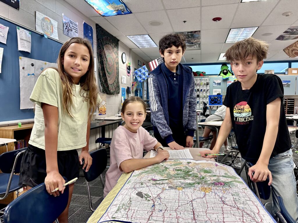 Four students collaborate at a table in a classroom, studying a large map and discussing their work.