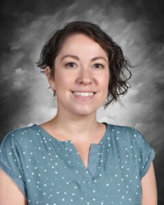 A smiling woman with short, curly hair wearing a blue polka dot shirt, set against a grey background.