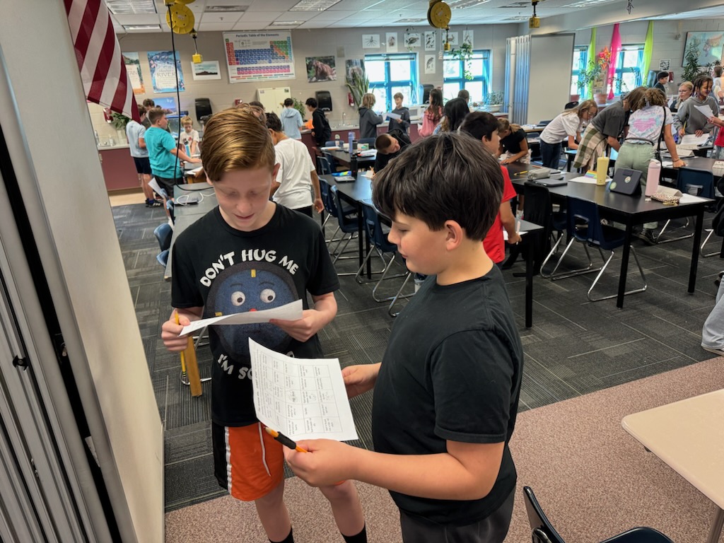 Two students stand in a classroom, discussing papers while other classmates work at nearby desks.