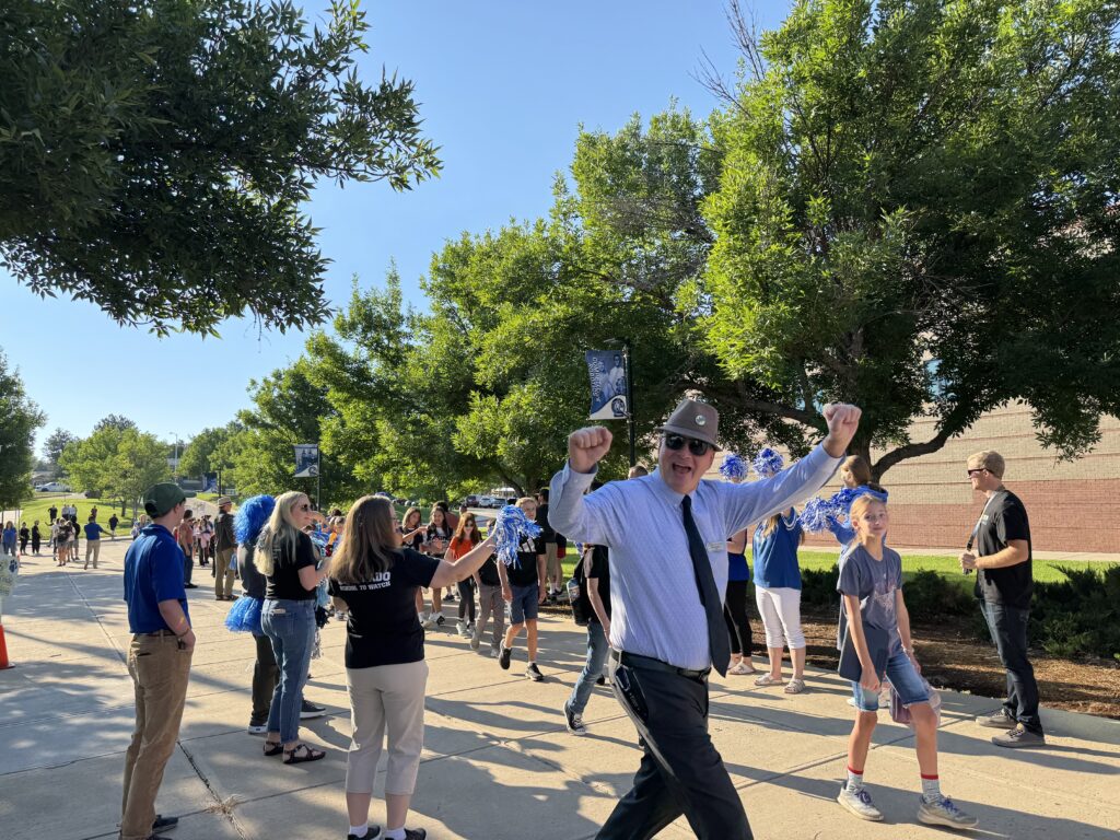 A lively scene with a crowd celebrating outdoors, featuring a man in a suit dancing and cheering among onlookers.