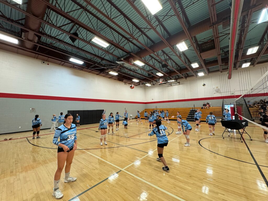 A volleyball practice session in a gym with players in blue jerseys, some preparing to serve and others in action.