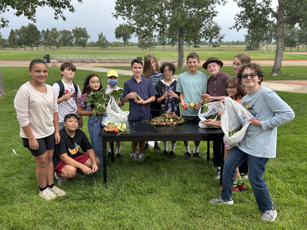 A group of children stands around a table with various vegetables, smiling and holding produce in a park setting.