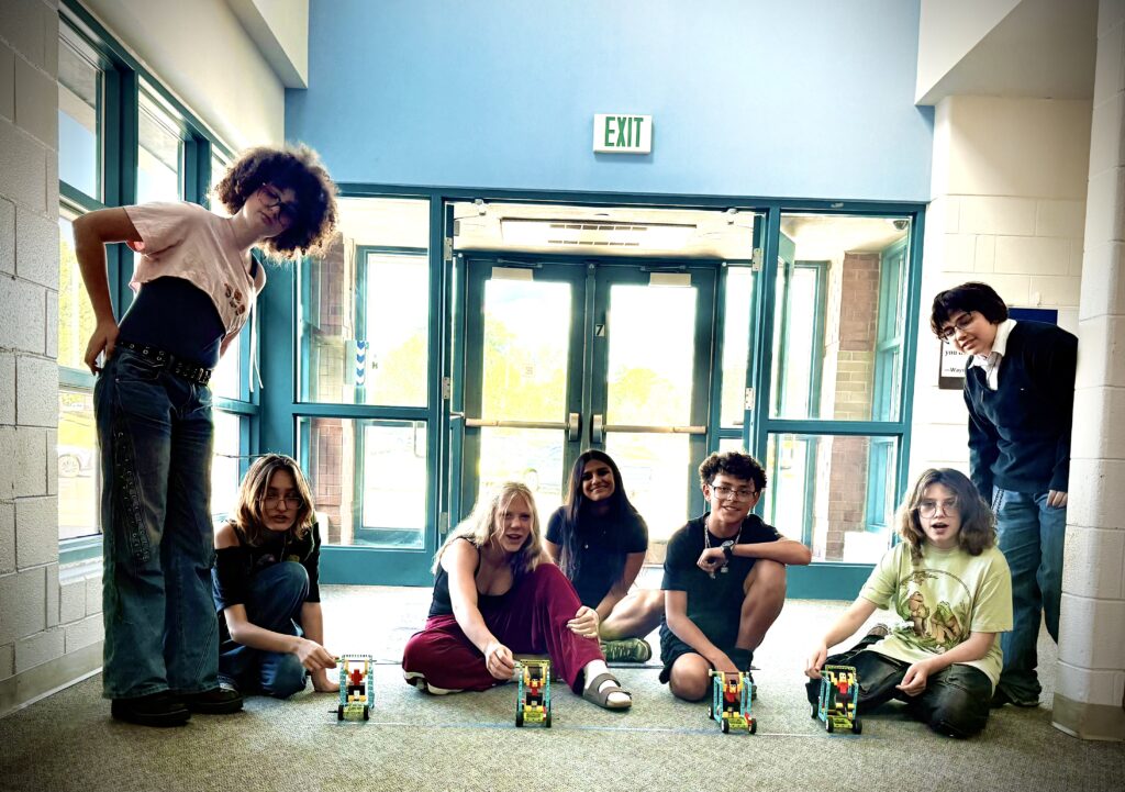 A group of seven young people poses indoors, displaying LEGO creations on the floor in front of them.