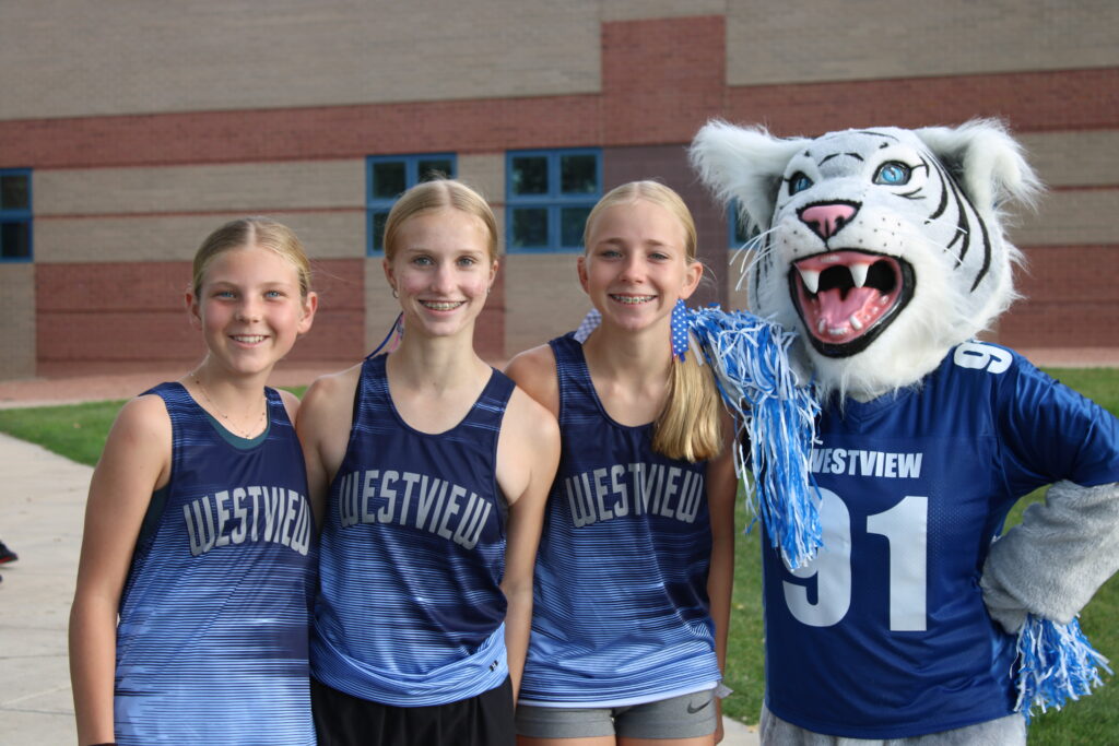 Three young athletes in Westview jerseys pose with a mascot in a blue and white tiger costume outside a school.